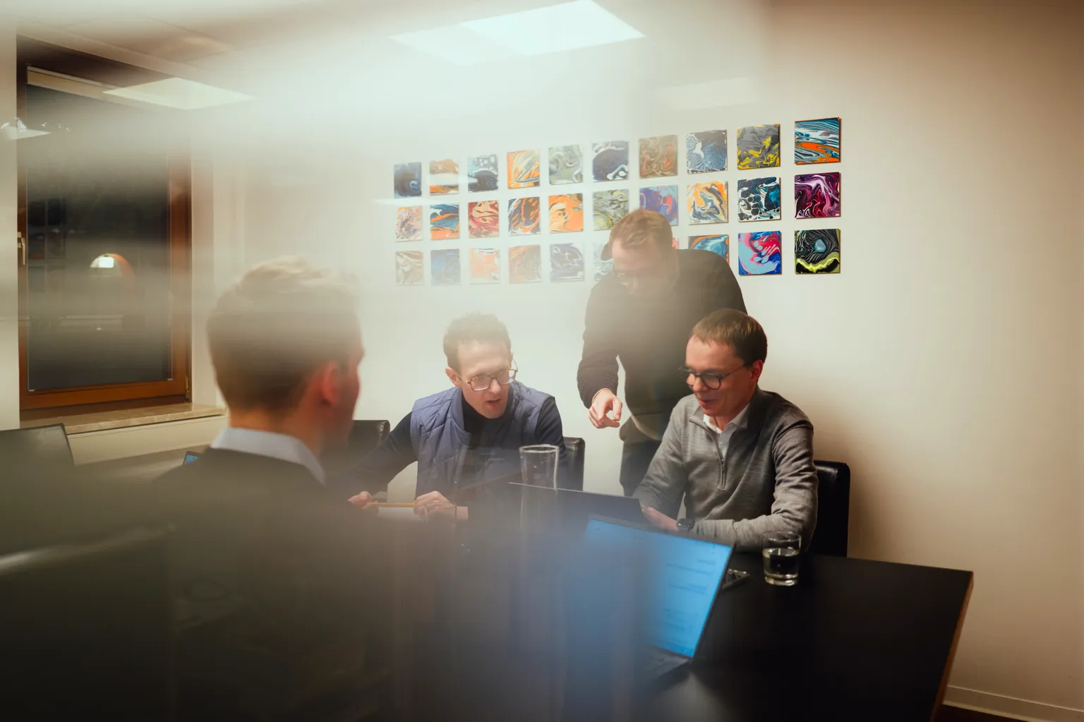 A motion-blurred candid photograph of three team members collaborating around a laptop in a modern office. The background features a gallery wall of small, colorful abstract art pieces.